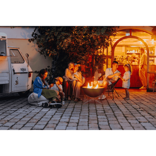 Höfats Bowl vuurschaal. Zwart geëmailleerd schaal met rvs onderstel. Brandend op een terras met diverse mensen eromheen.