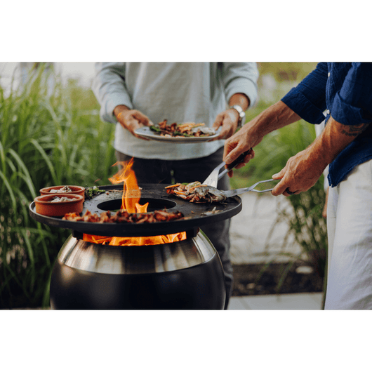 Höfats MOON 45 vuurkorf roestvrijstaal met hoge roestvrijstalen standaard. Voorzien van Geëmailleerd gietijzeren grillring en brandend vuur op een terras – rookvrije design vuurkorf met houtvergassingstechnologie