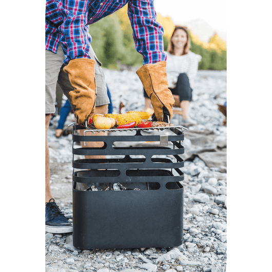 Zomerse setting met man die het grillrooster met etenswaar op de  Höfats Cube vuurkorf zet. Vrouw kijkt op de achtergrond naar het tafereel.
