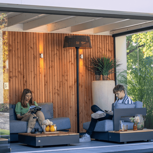 Zwarte staande heater met olifin kap bij een loungeset onder houten veranda met glazen deur. 